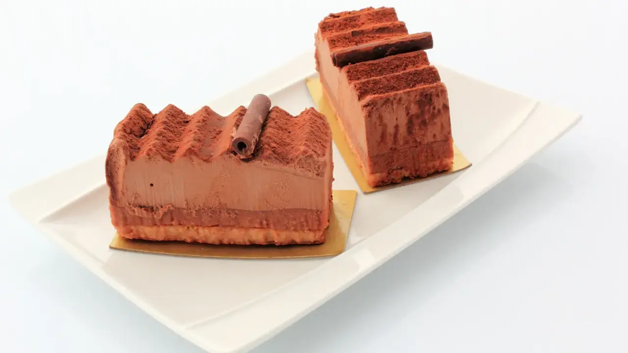 Deux parts de gâteau Trianon, le meilleur pâtissier, saupoudrées de cacao, sur une assiette blanche.