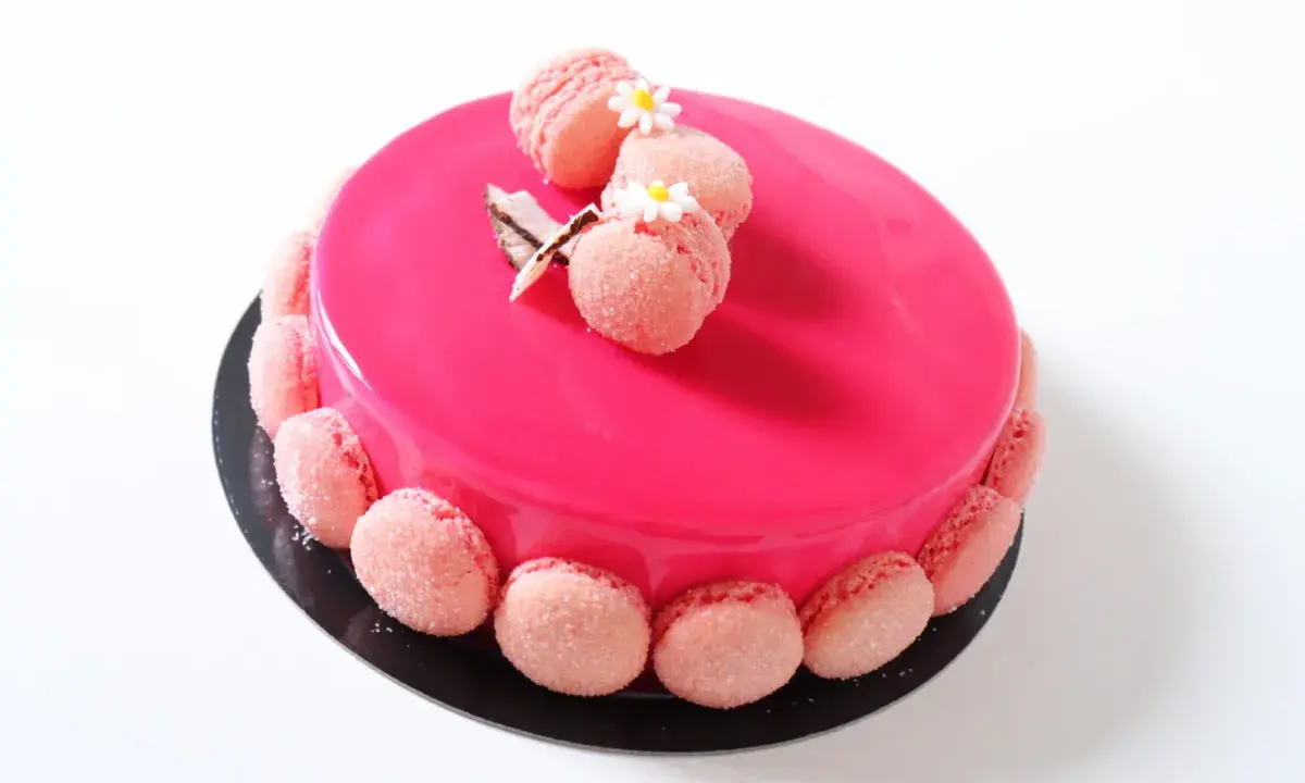 Entremet fraise rose vif, décoré de macarons roses et de petites fleurs blanches.