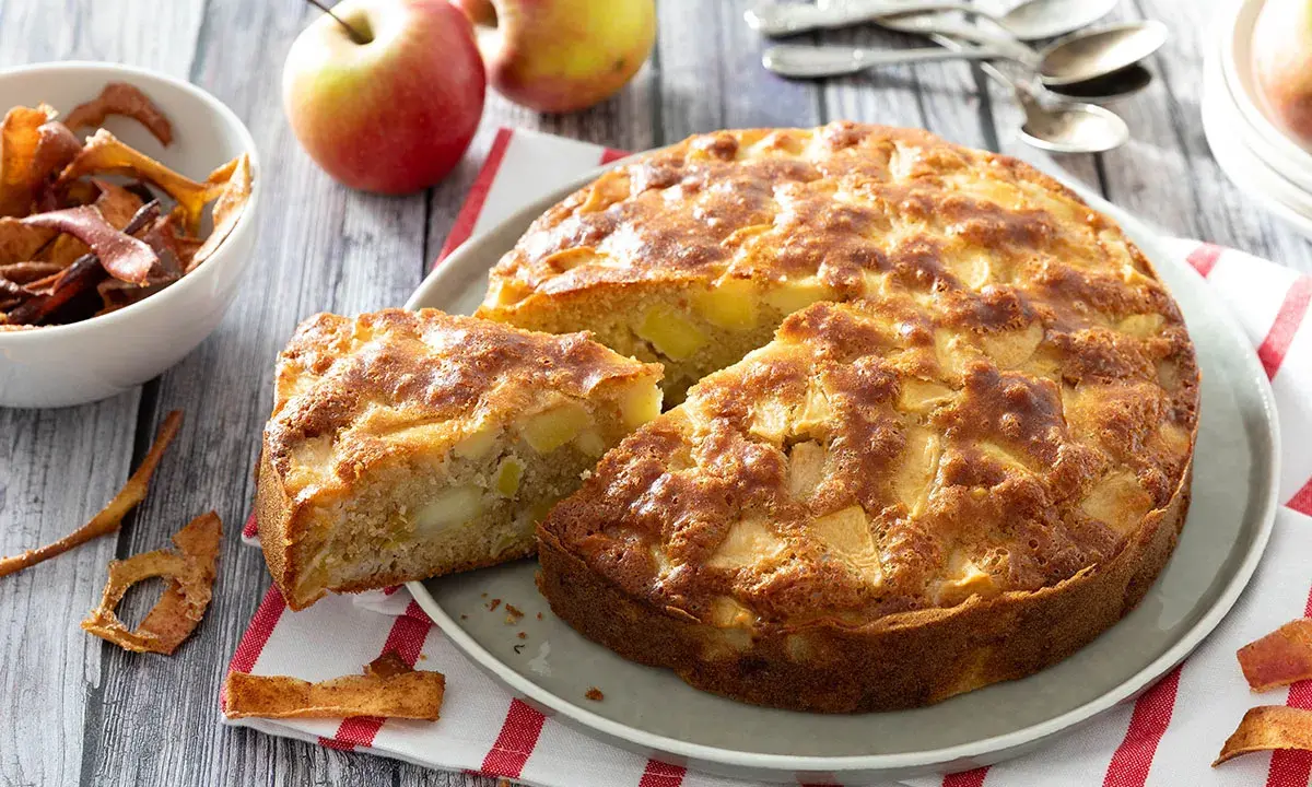 Un gâteau aux pommes super moelleux, doré et appétissant, avec des morceaux de pommes visibles. Des pommes fraîches et des chips de pommes sont disposées autour.