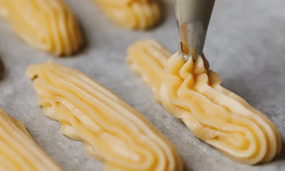 Préparation de la pâte à choux pour la recette de maman choux à la crème. La poche à douille dépose des éclairs sur du papier cuisson.