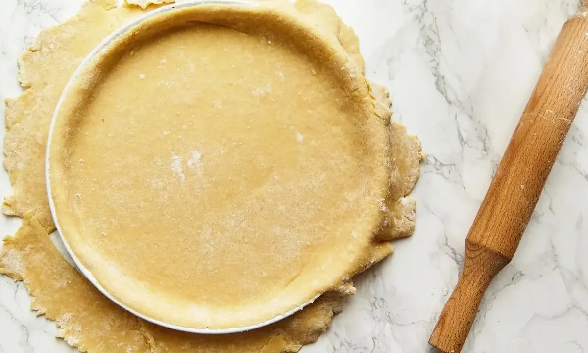Pâte sablée au robot prête à être garnie, posée dans un moule à tarte. Rouleau à pâtisserie à côté.