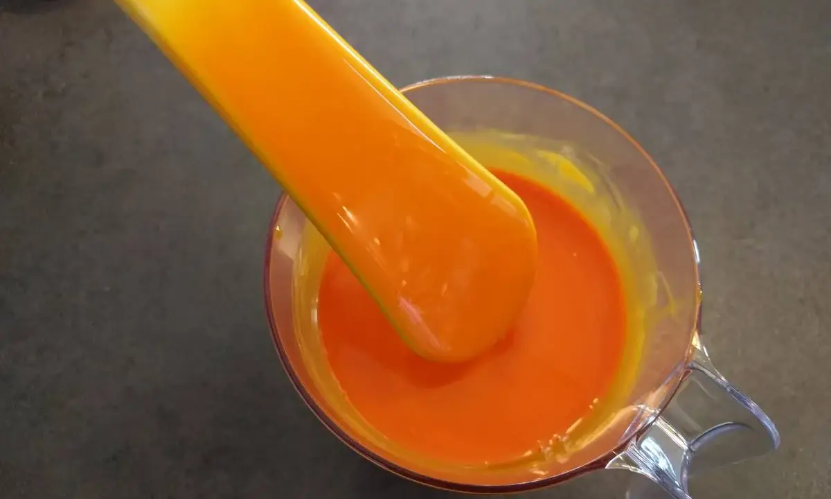 Une spatule verse un glaçage miroir orange vif dans un récipient en verre.