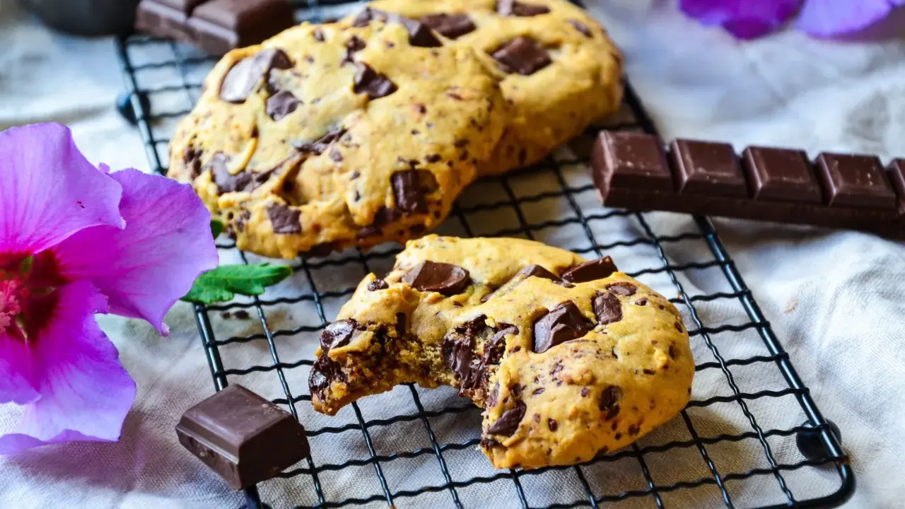 Gros plan sur des cookies chocolat faits maison, un est croqué, révélant un cœur fondant. Des morceaux de chocolat et une fleur d'hibiscus violette complètent la scène.