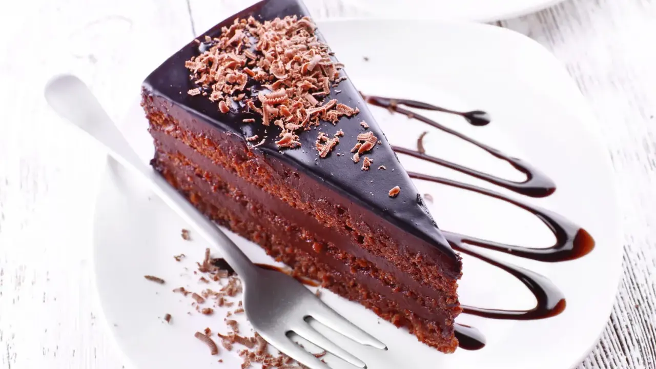 Une part de gâteau aux chocolats, nappée de glaçage brillant et garnie de copeaux de chocolat, est servie sur une assiette blanche avec un filet de sauce chocolat.