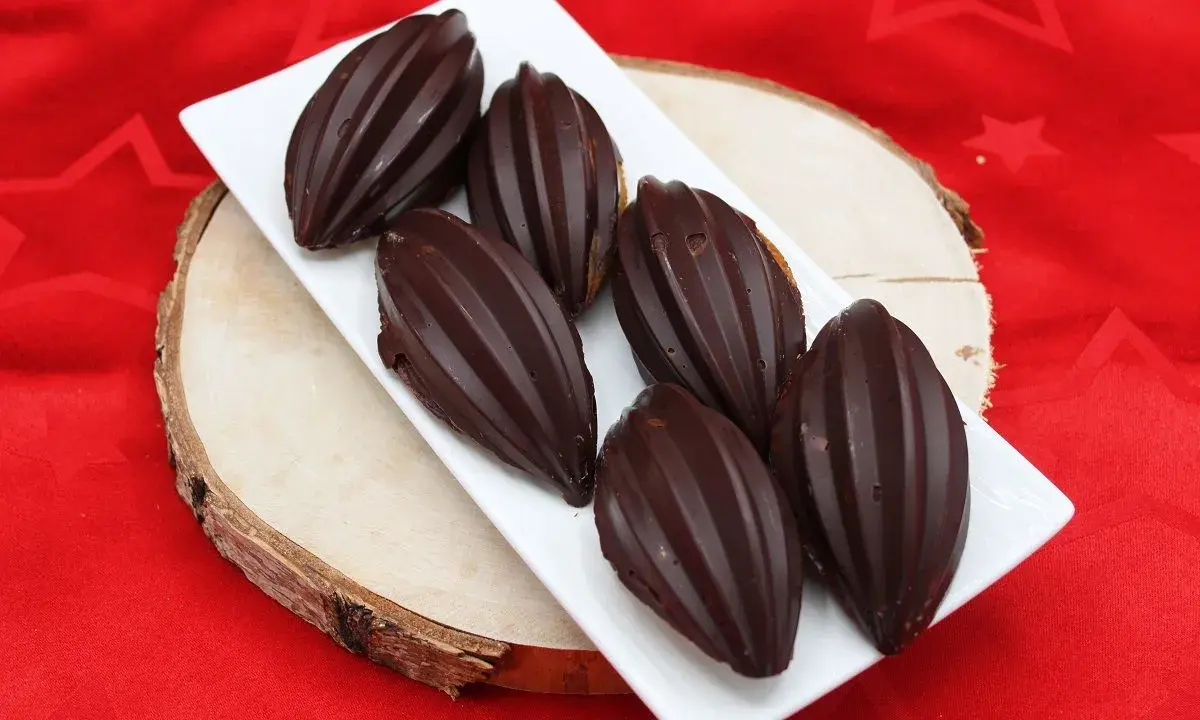 Six financiers chocolat, en forme de fèves de cacao, sur une assiette blanche, posée sur une planche de bois, sur un tissu rouge étoilé.