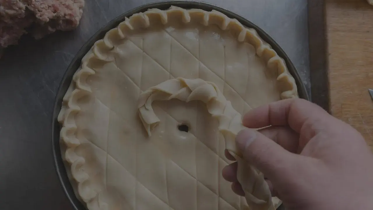 Main d'un pâtissier décorant une tarte avec une pâte brisée tressée. Un tas de farce est visible à gauche.