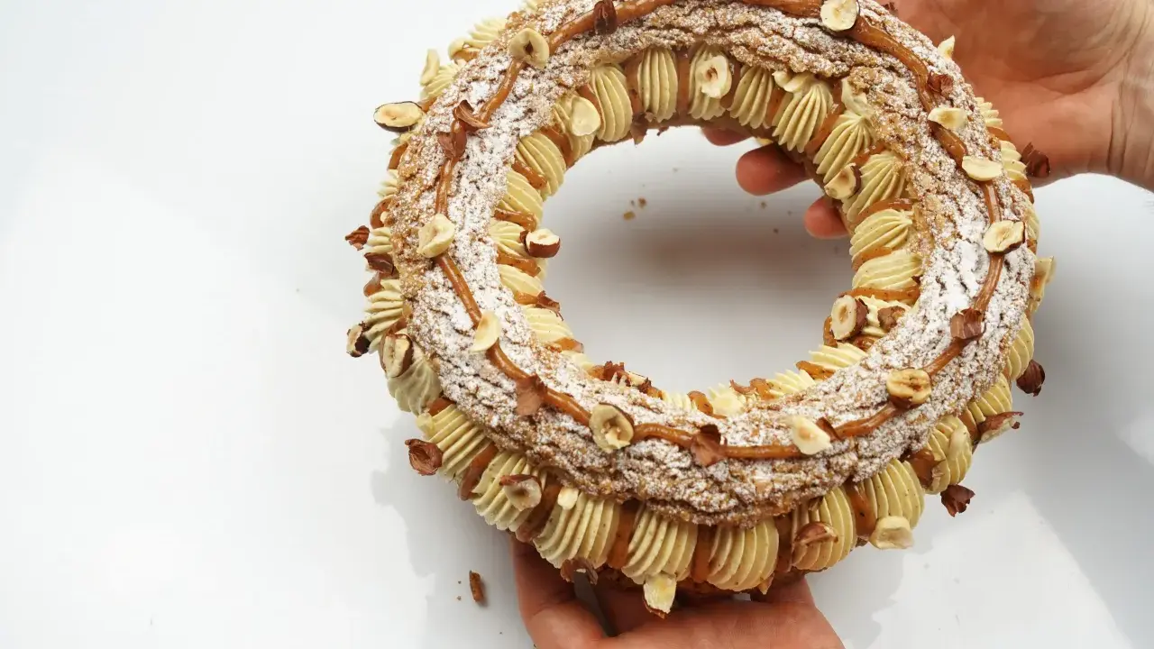 Un Paris-Brest craquelin, orné de noisettes et de caramel, est tenu par des mains.