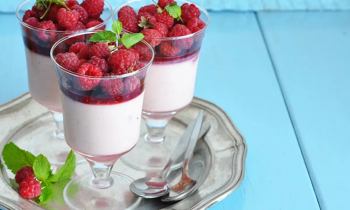 Trois panna cotta onctueuses, garnies de framboises fraîches et d'un coulis de framboise, sur un plateau argenté.