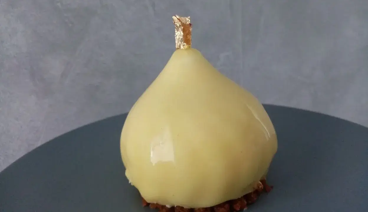 Délice en forme de poire, recouvert d'un glacage miroir jaune éclatant, sur un lit de crumble croquant, orné d'une touche de feuille d'or.