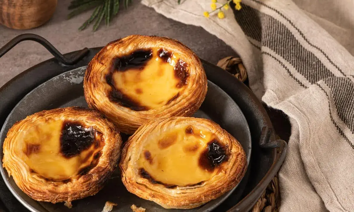 Trois délicieux pasteis de nata dorés, leur crème onctueuse légèrement caramélisée, sur un plateau en métal.