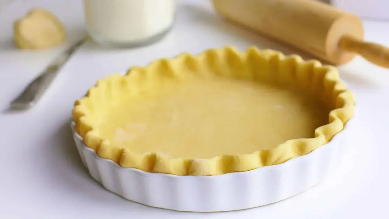 Pâte brisée prête pour la cuisson dans un plat à tarte blanc, avec un rouleau à pâtisserie en arrière-plan.