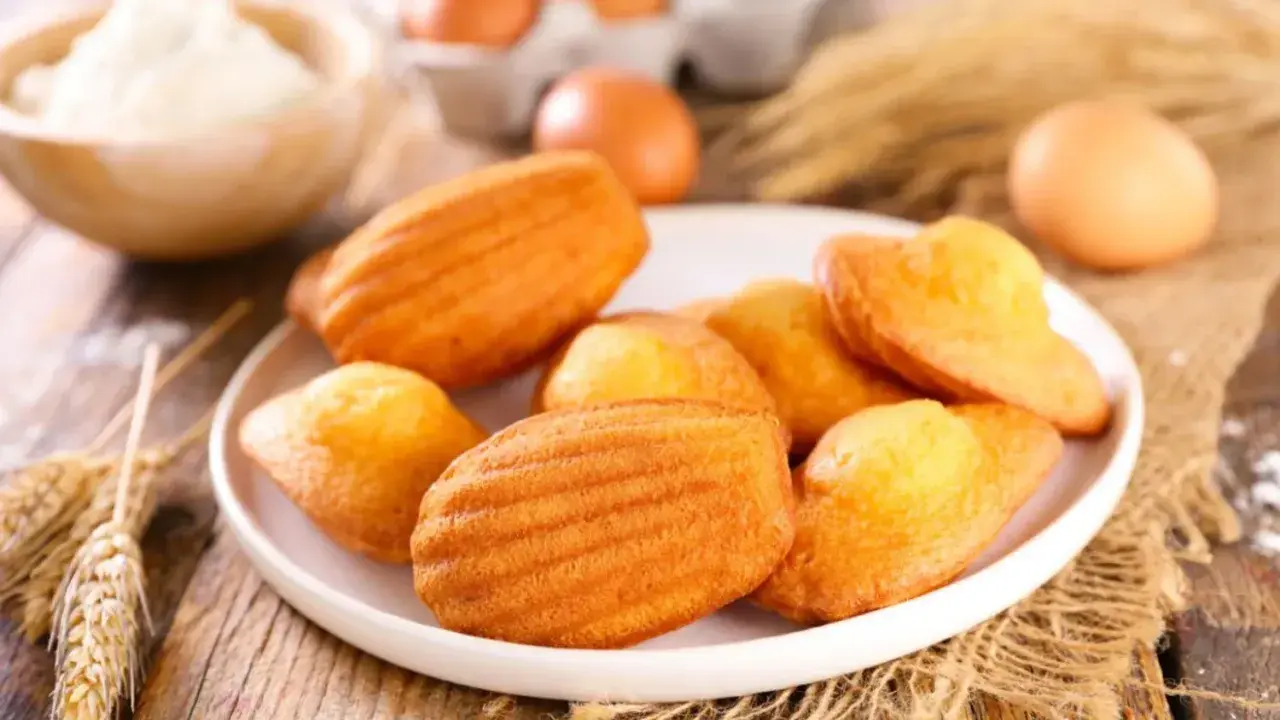 Des madeleines dorées, parfaites pour une recette gâteau madeleine meilleur pâtissier. Des œufs et de la farine en arrière-plan.