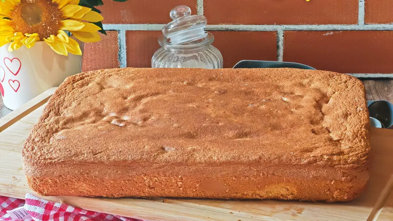 Une génoise rectangulaire dorée, parfaite pour 15 personnes, attend d'être garnie. Des fleurs jaunes et un fond de briques rouges créent une ambiance chaleureuse.