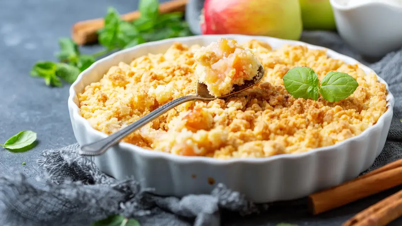 Un délicieux crumble aux pommes à l'ancienne, garni de feuilles de menthe fraîche, avec des pommes et des bâtons de cannelle en arrière-plan.