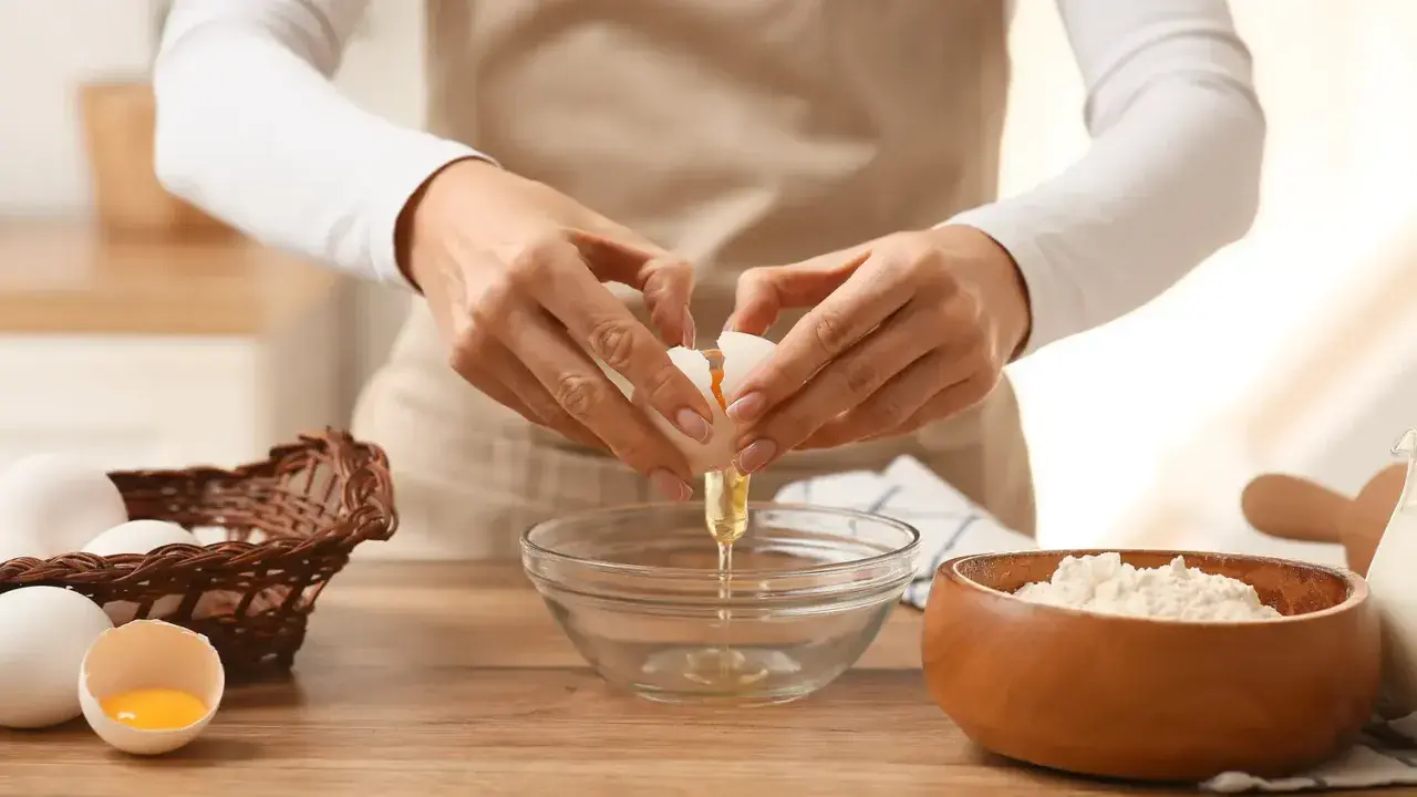 Préparation d'une recette pâte brisée facile : une main casse un œuf dans un bol, à côté d'un panier d'œufs et d'un bol de farine.