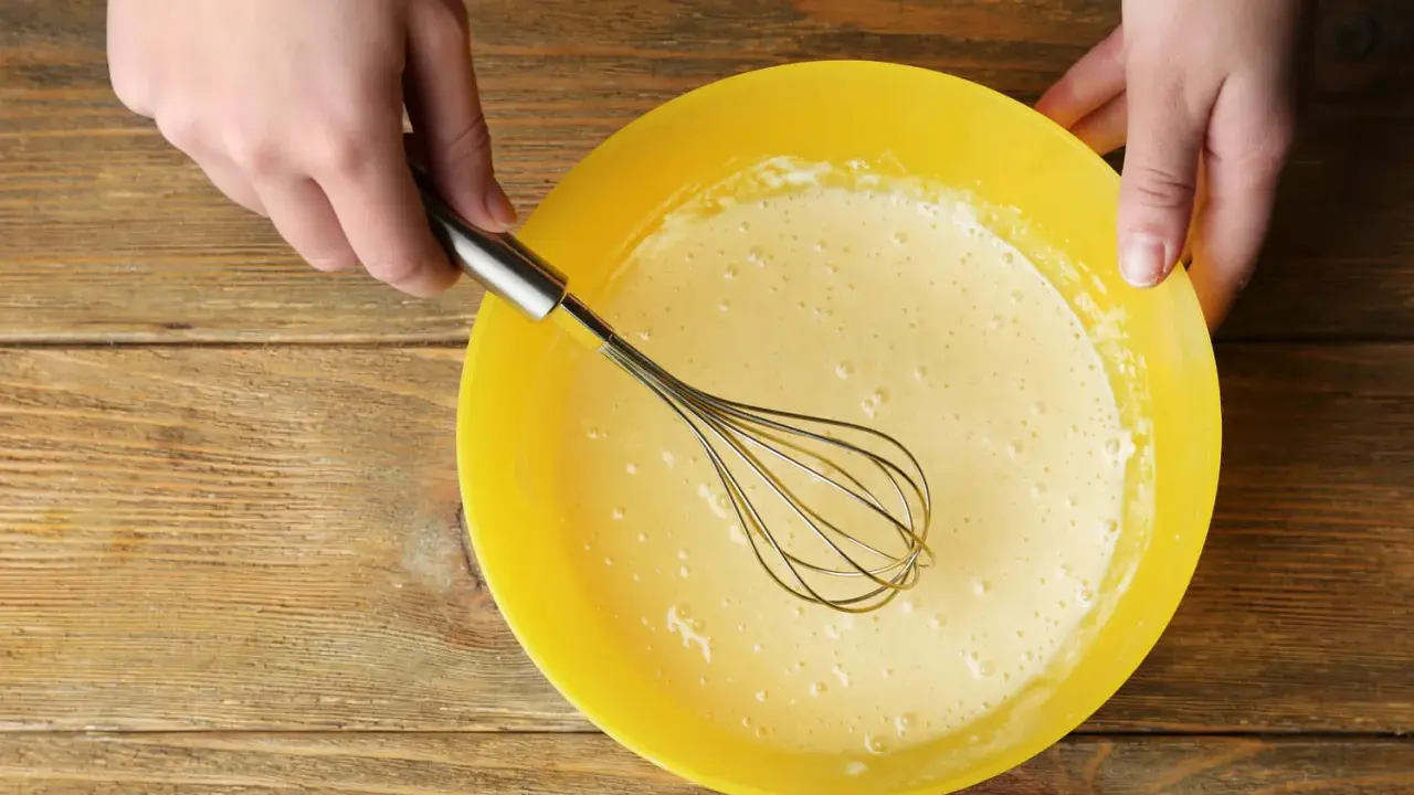 Mains préparant la pâte à crêpes dans un bol jaune. La pâte est prête pour son temps de repos.