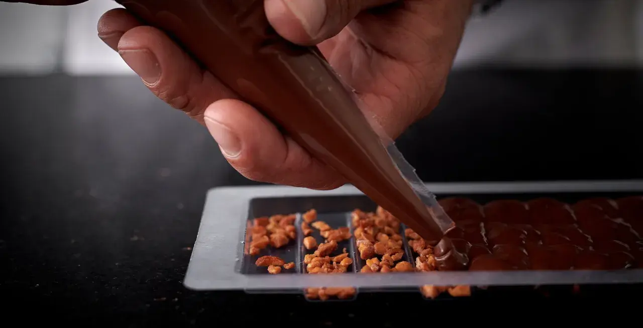 Main d'un artisan versant du chocolat fondu dans un moule pour créer une tablette de chocolat maison garnie de noisettes caramélisées.