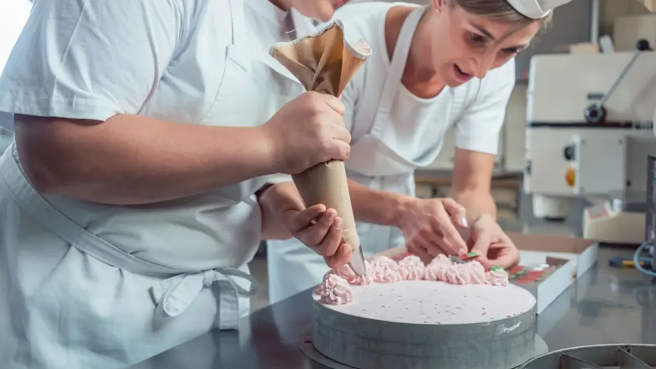 Deux pâtissières décorent un gâteau rose lors d'un cours de pâtisserie en ligne. L'une utilise une poche à douille, l'autre des fruits.