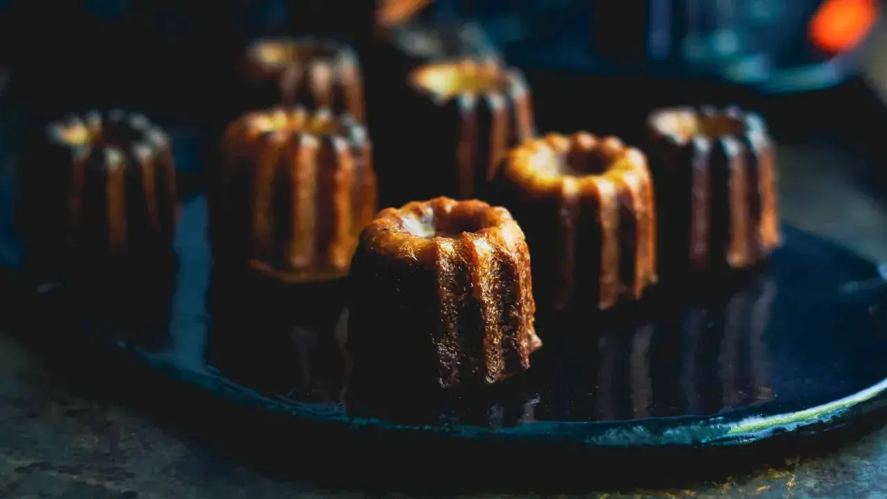 Plusieurs cannelés dorés, parfaits pour la conservation, sont disposés sur une assiette sombre.