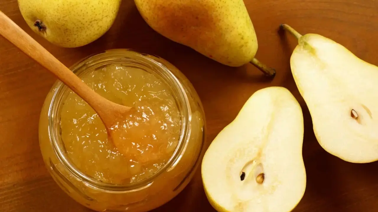 Un pot de confiture de poires maison, avec une cuillère en bois, entouré de poires fraîches coupées et entières sur fond de bois.