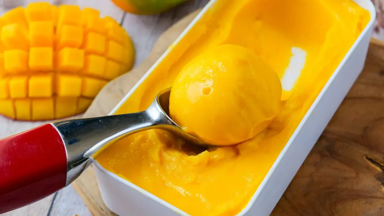 Une cuillère prélève une boule de sorbet mangue crémeux d'une boîte. Des morceaux de mangue fraîche sont visibles à côté.