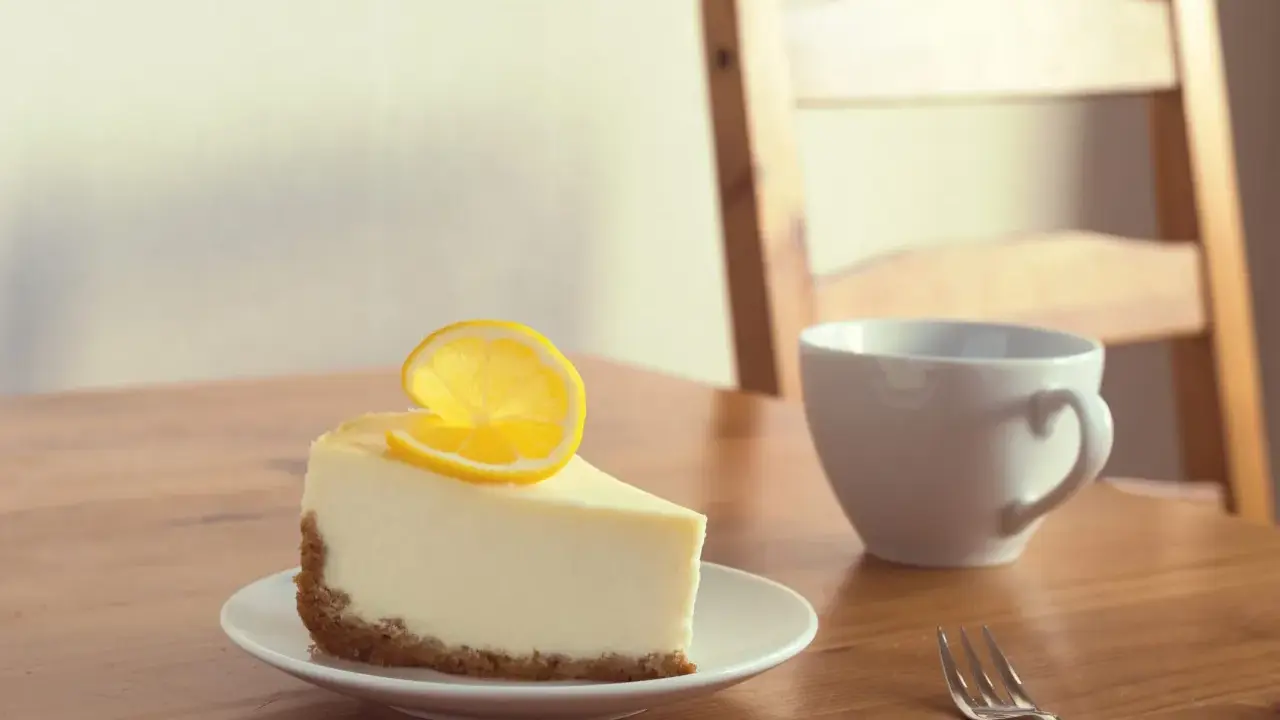 Une part de le meilleur cheesecake au citron, garnie d'une tranche de citron, sur une assiette blanche, à côté d'une tasse et d'une fourchette sur une table en bois.