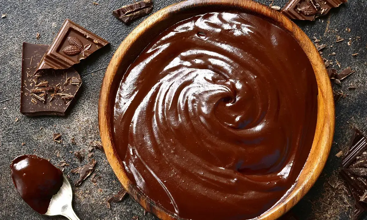 Un bol en bois déborde de chocolat fondu, prêt à faire du chocolat. Des éclats de chocolat noir et une cuillère en argent complètent la scène gourmande.