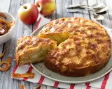Un gâteau aux pommes super moelleux, doré et appétissant, avec des morceaux de pommes visibles. Des pommes fraîches et des chips de pommes sont disposées autour.