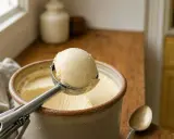 Une cuillère prélève une boule crémeuse de glace vanille maison dans un pot en grès.