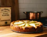 Tarte aux poires au sirop, dorée et appétissante, sur un plan de travail en bois. Un pot en cuivre est visible en arrière-plan.