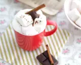 Une tasse rouge débordant de guimauves et de chocolat, parfaite pour une recette guimauve chocolat réconfortante. Un bâton de cannelle et des carrés de chocolat complètent cette gourmandise.