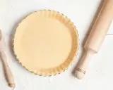 Pâte à tarte crue dans un moule, prête pour le temps de cuisson. Rouleau à pâtisserie et cuillère en bois à proximité.