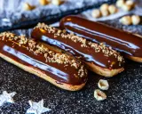 Trois éclairs au chocolat brillants, saupoudrés de noisettes concassées, sur un fond sombre parsemé de sucre glace.
