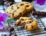 Gros plan sur des cookies chocolat faits maison, un est croqué, révélant un cœur fondant. Des morceaux de chocolat et une fleur d'hibiscus violette complètent la scène.