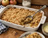 Un plat de meilleur crumble aux pommes tout juste sorti du four, avec une cuillère prête à servir. Des pommes fraîches et une crème anglaise complètent ce délice.