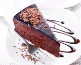 Une part de gâteau aux chocolats, nappée de glaçage brillant et garnie de copeaux de chocolat, est servie sur une assiette blanche avec un filet de sauce chocolat.