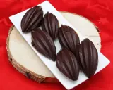 Six financiers chocolat, en forme de fèves de cacao, sur une assiette blanche, posée sur une planche de bois, sur un tissu rouge étoilé.