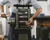 Une personne utilise un laminoir manuel pour travailler le métal. Les rouleaux sont prêts à aplatir un fil.