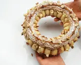 Un Paris-Brest craquelin, orné de noisettes et de caramel, est tenu par des mains.