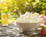 Chantilly aérienne dans un bol, fouet, fraises et limonade sur une table en bois ensoleillée.
