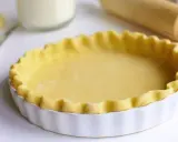 Pâte brisée prête pour la cuisson dans un plat à tarte blanc, avec un rouleau à pâtisserie en arrière-plan.