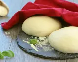 Deux boules de pâte à pizza professionnelle prêtes à être travaillées, sous un torchon rouge. Une feuille de basilic et une cuillère de semoule complètent la scène.