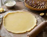 Pâte à tarte prête à être utilisée, avec un rouleau à pâtisserie et des prunes. Le fond de tarte croustillant attend sa garniture.