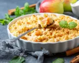 Un délicieux crumble aux pommes à l'ancienne, garni de feuilles de menthe fraîche, avec des pommes et des bâtons de cannelle en arrière-plan.