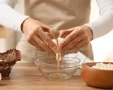 Préparation d'une recette pâte brisée facile : une main casse un œuf dans un bol, à côté d'un panier d'œufs et d'un bol de farine.