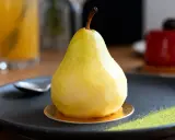 Un entremet poire, glacé et brillant, repose sur une assiette noire avec une touche de poudre verte.