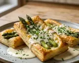 Trois feuilletés aux asperges, dorés et garnis de fromage fondant, de ciboulette et de sauce crémeuse, sur une assiette grise.