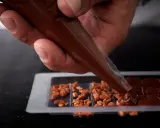 Main d'un artisan versant du chocolat fondu dans un moule pour créer une tablette de chocolat maison garnie de noisettes caramélisées.