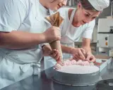Deux pâtissières décorent un gâteau rose lors d'un cours de pâtisserie en ligne. L'une utilise une poche à douille, l'autre des fruits.