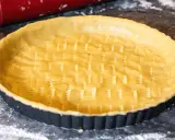 Un fond de tarte amande prêt à être garni, avec un rouleau à pâtisserie rouge et un œuf à proximité.
